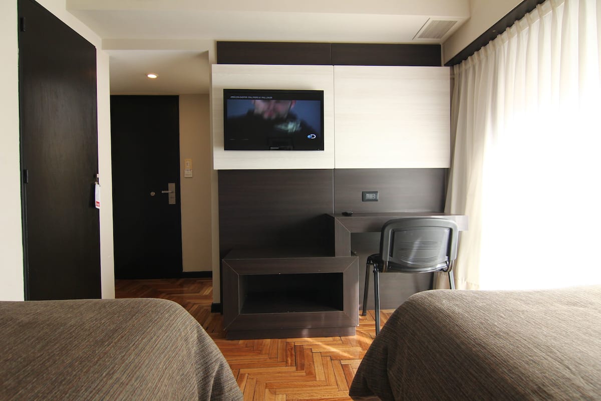 Habitación de hotel con dos camas, un televisor montado en la pared, un escritorio y una silla.