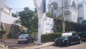 Una strada con auto parcheggiate, un edificio bianco con ingresso ad arco e un cartello pubblicitario con scritto 'Sherezada'.