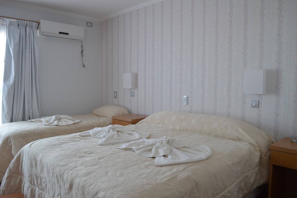 A hotel room with two beds, a window with curtains, an air conditioning unit, and a wall-mounted light fixture.