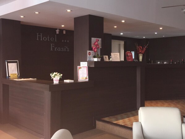A hotel reception area with a dark reception desk, a white chair, and a carpeted floor. - Hotel Frasis (San Clemente)