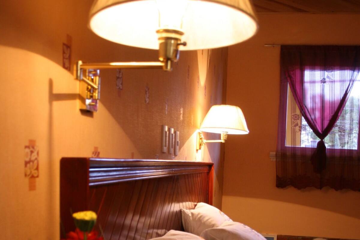 A hotel room with a bed, bedside lamps, a nightstand with a flower vase, and a window with curtains.
