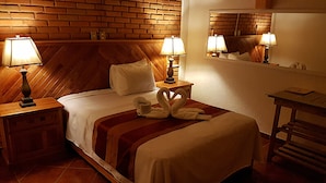 Een hotelkamer met een bakstenen muur, een houten hoofdeinde, twee nachtlampjes, een bed met een witte deken, een rood-wit gestreepte plaid en een spiegel aan de muur.