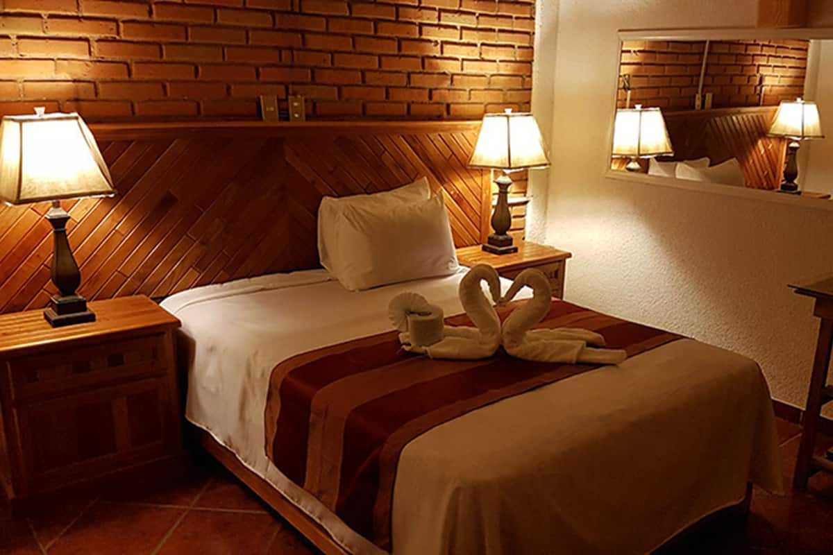A hotel room with a brick wall, wooden headboard, two bedside lamps, a bed with a white comforter, a red and white striped blanket, and a mirror on the wall.