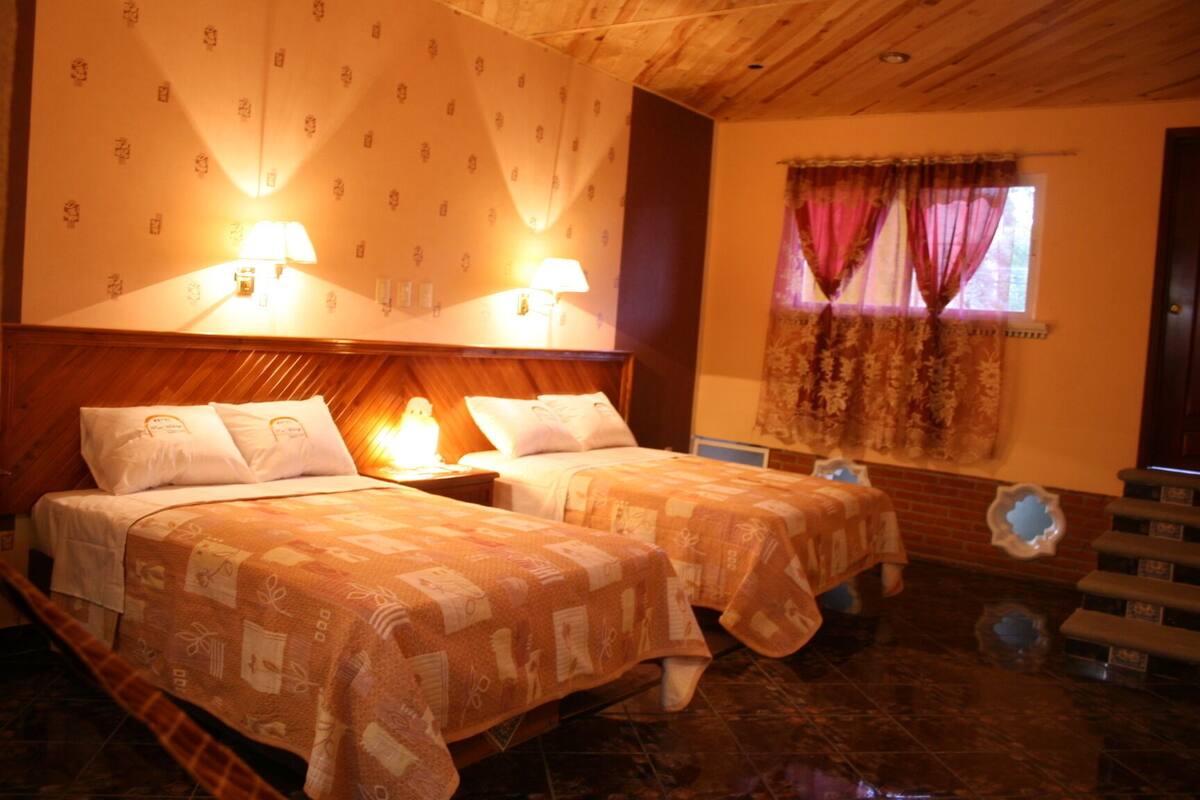 A hotel room with two beds, wooden headboard, patterned wallpaper, two bedside lamps, a window with curtains, and a small decorative item on the floor.