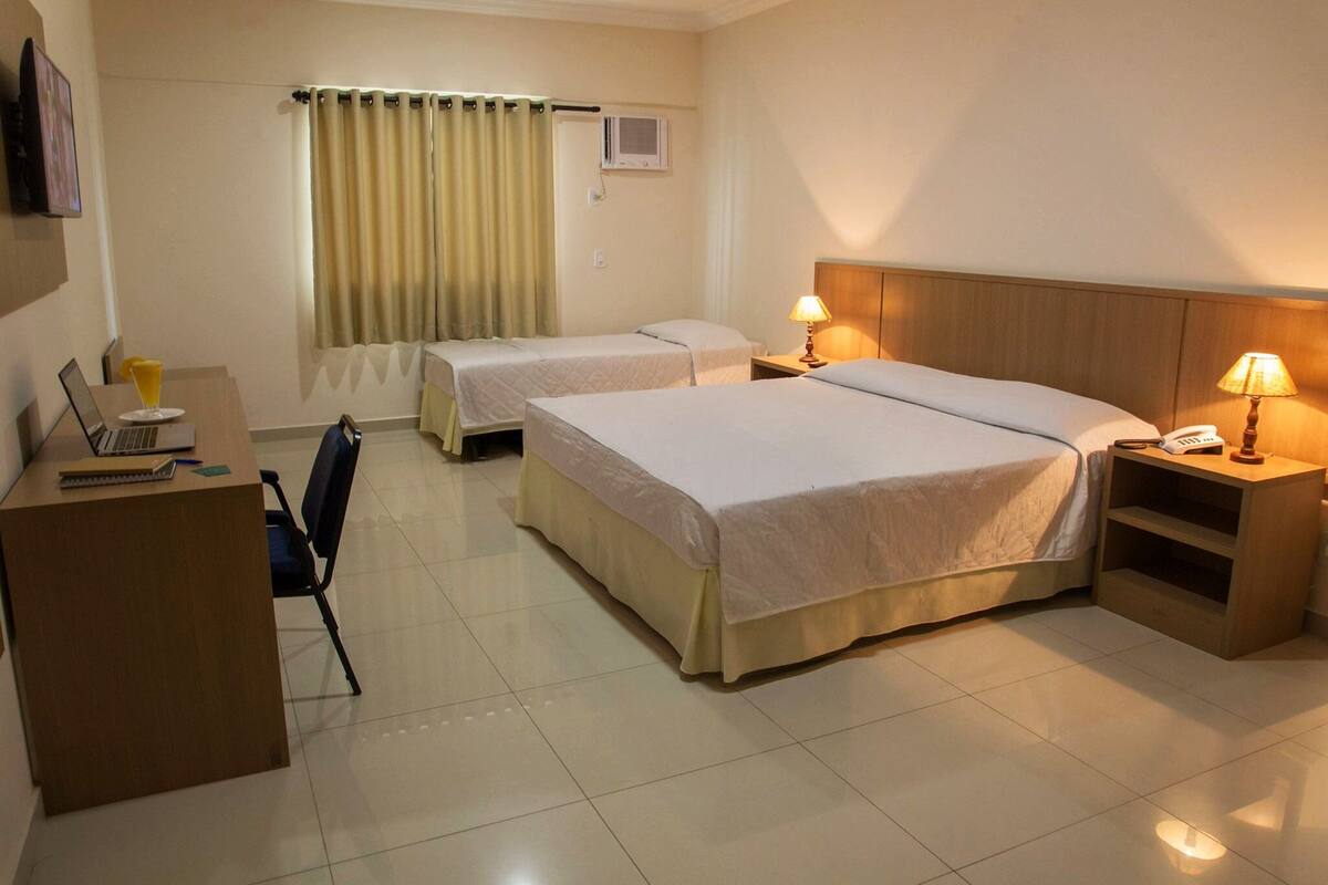 Een hotelkamer met twee bedden, een bureau met een laptop, een stoel, een nachtkastje met een lamp en een raam met gordijnen.