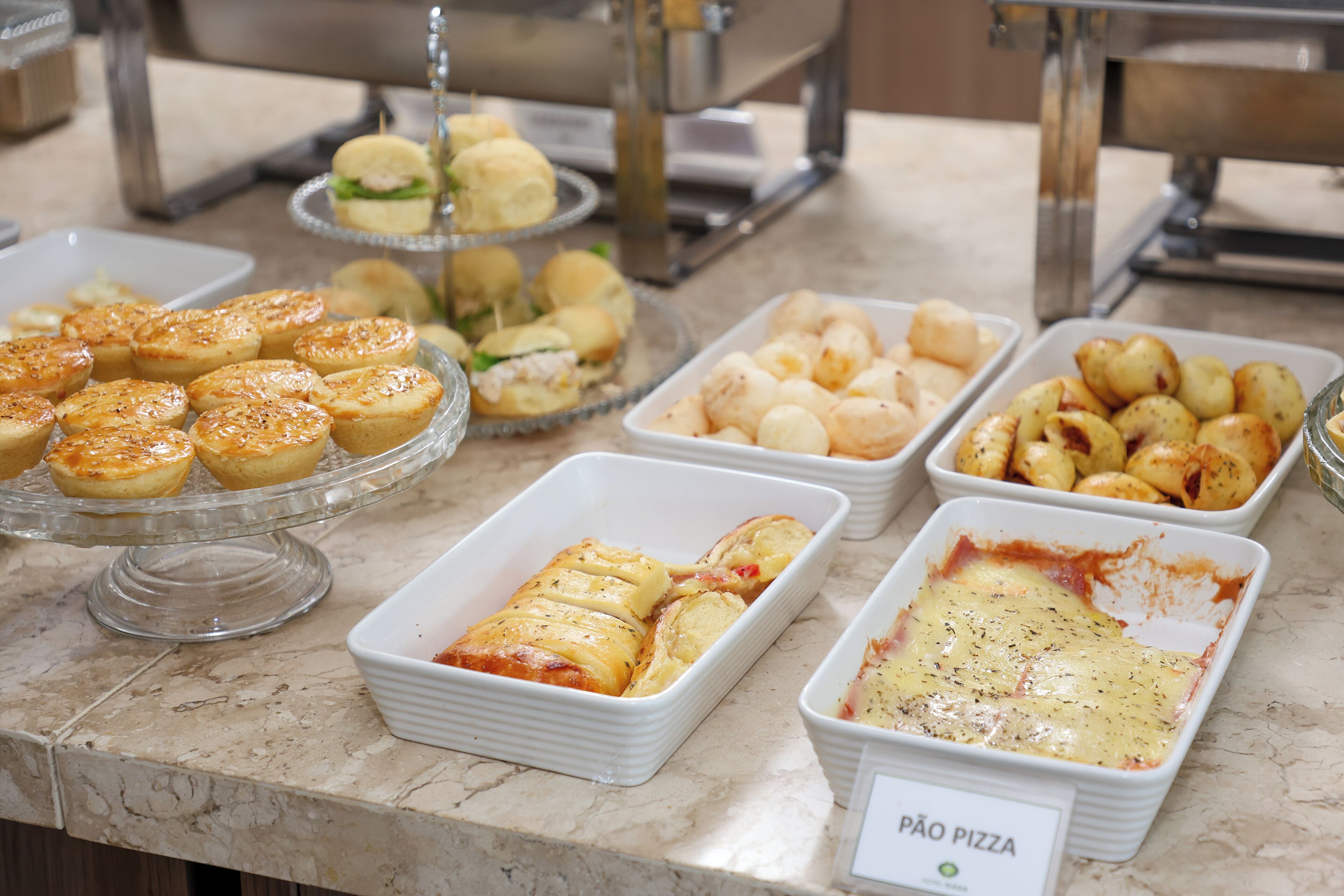 Un bufé con diversas opciones de panadería, incluyendo pasteles, pan y un plato etiquetado como 'Pão Pizza'.
