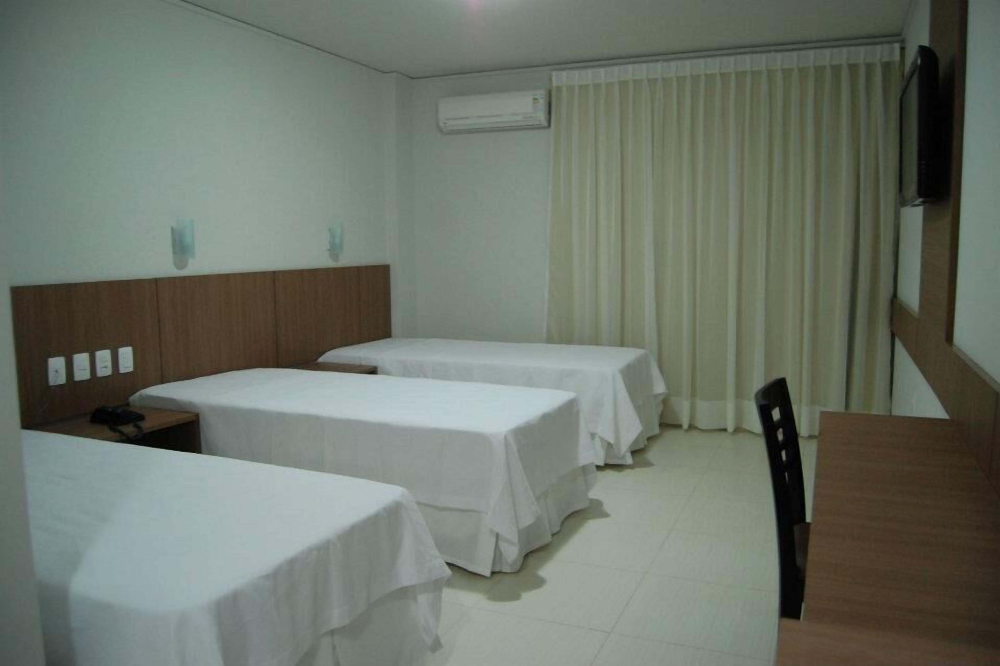 A hotel room with three single beds, a wooden headboard, a desk with a chair, and a television mounted on the wall.