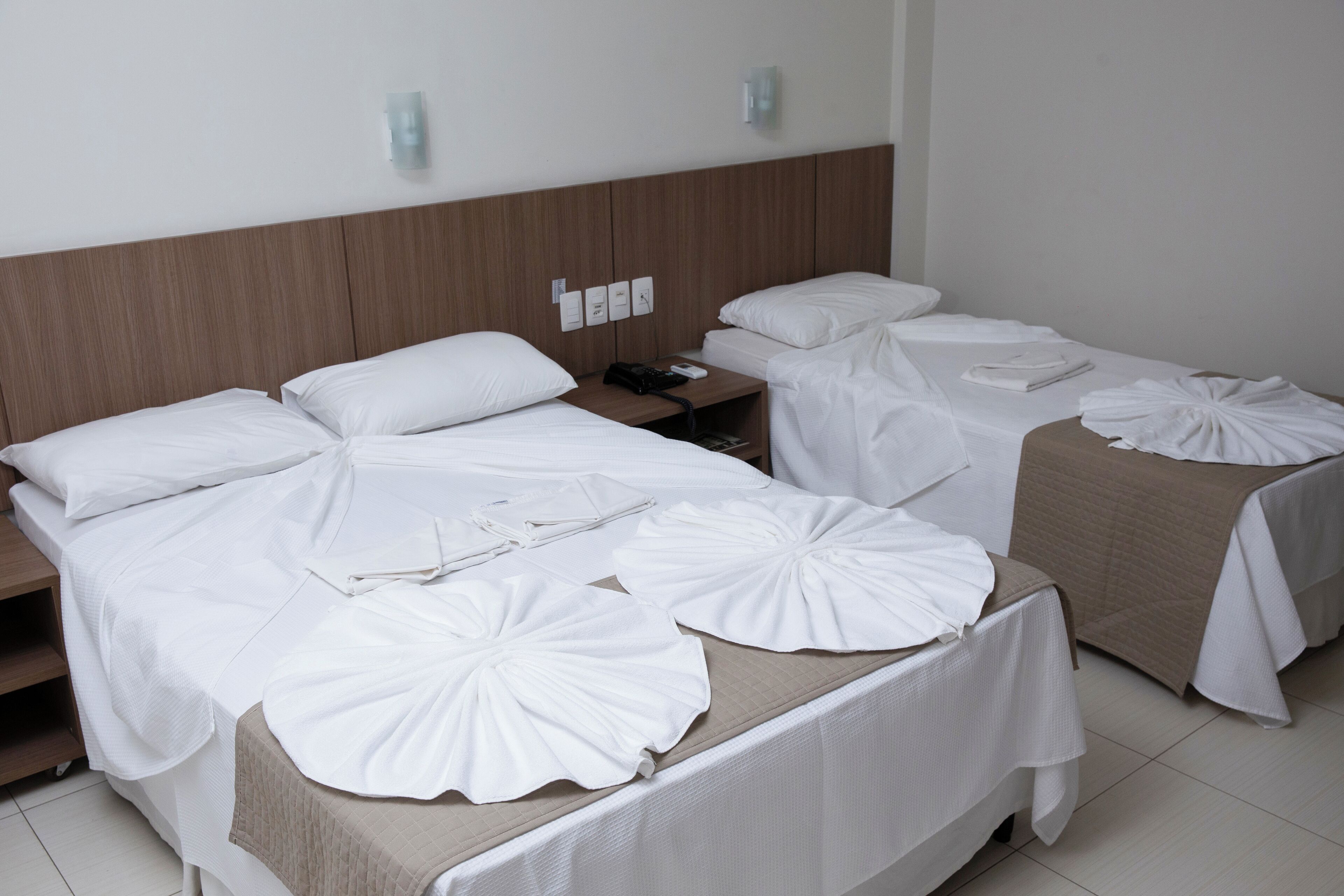 A hotel room with two beds, each with white bedding and decorative pillows. There is a wooden headboard, a nightstand with a phone and a lamp, and a wall-mounted light fixture.