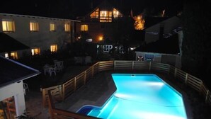 Una piscina con plataforma de madera y valla, rodeada de edificios con ventanas iluminadas.