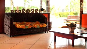 A cozy living area with a wicker sofa, a wooden coffee table, and a view of a garden through large windows. - Água Doce Praia Hotel (Ubatuba)