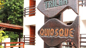 A wooden sign with directional arrows pointing to 'TRILHA,' 'QUIOSQUE,' 'LAGO,' and 'MIRANTE.' - Água Doce Praia Hotel (Ubatuba)