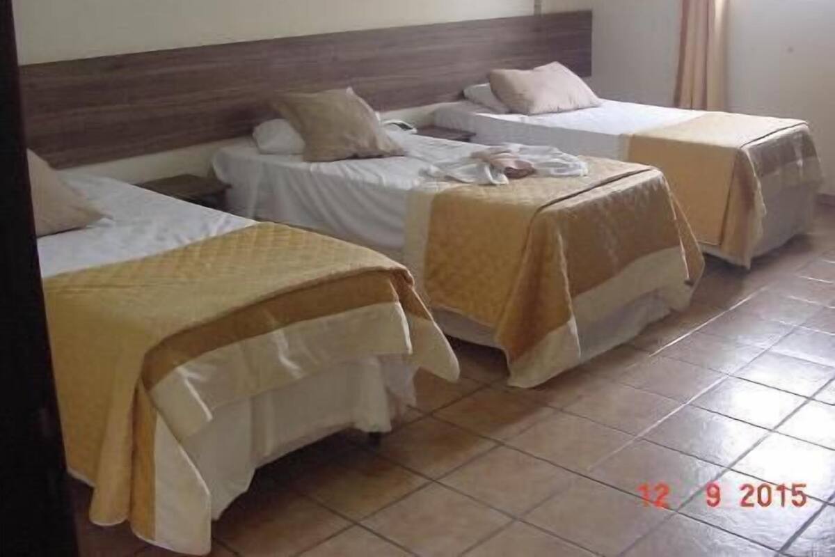 Une chambre d’hôtel avec trois lits, chacun ayant un style de literie différent. La chambre est dotée d’un sol carrelé et d’une tête de lit en bois.