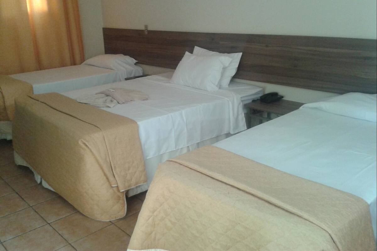 Une chambre d’hôtel avec trois lits, chacun recouvert d’une couverture beige. On y trouve une tête de lit en bois, une table de chevet avec un téléphone et un rideau sur le côté gauche.