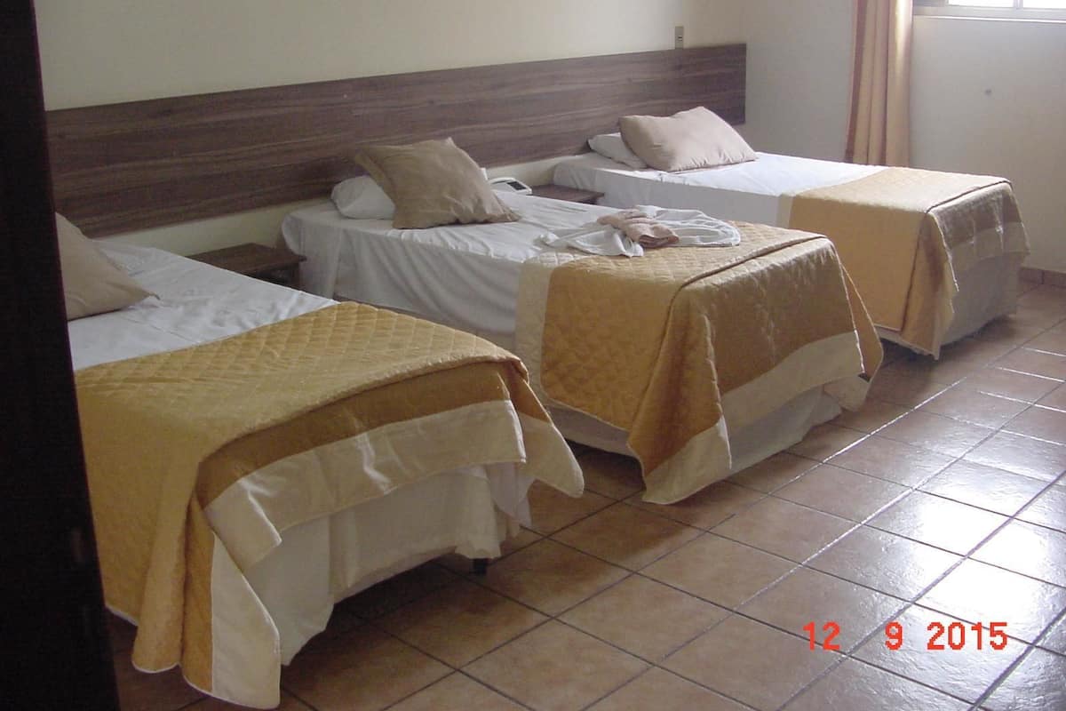 Une chambre d’hôtel avec trois lits simples, chacun étant  équipé d’une couverture et d’un oreiller de couleur beige. La chambre a un sol carrelé et une tête de lit en bois.