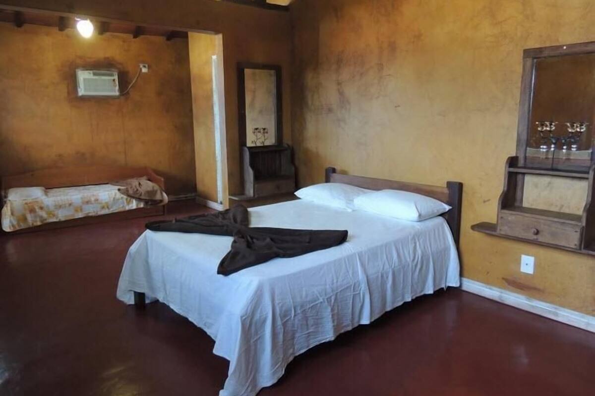 Une chambre avec un lit simple, un banc en bois, un radiateur fixé au mur et un miroir.