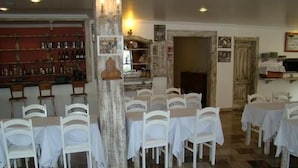Un restaurante con sillas y mesas blancas, una zona de bar con estantes llenos de botellas y un pilar de madera rústica.