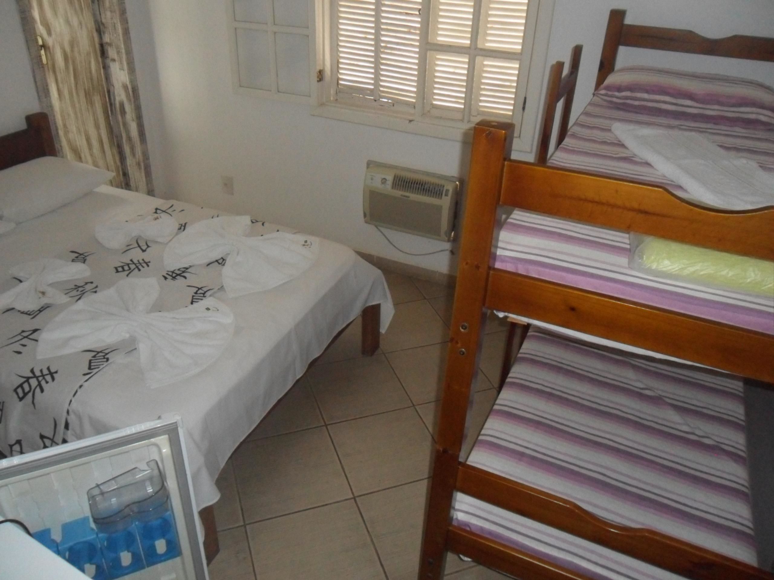 Un dormitorio con cama, literas de madera, ventana con persianas y un calentador fijado a la pared.