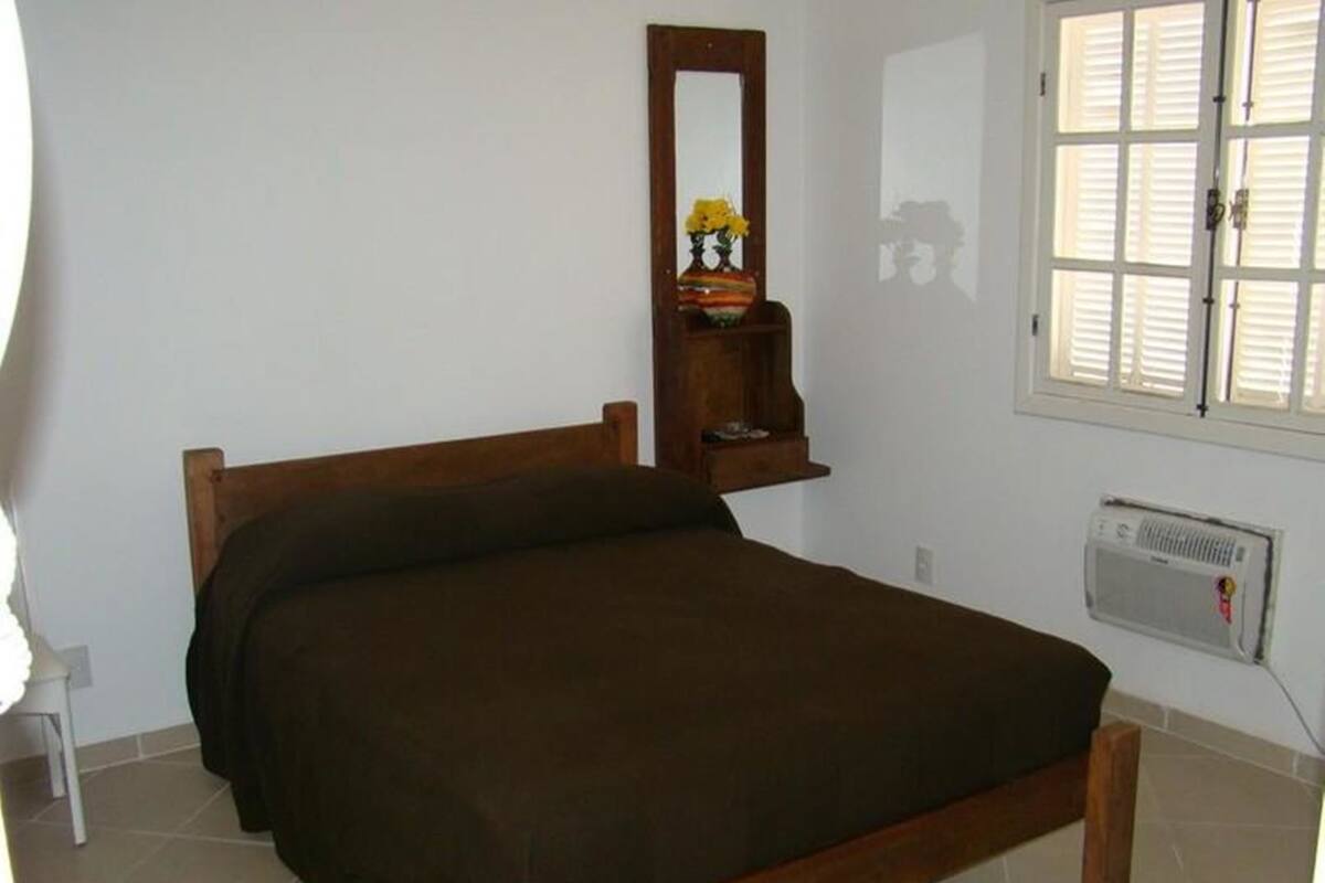 Un lit simple avec un cadre en bois et une couvre-lit marron foncé, dans une pièce aux murs blancs, avec une fenêtre équipée de stores et un radiateur fixé au mur.