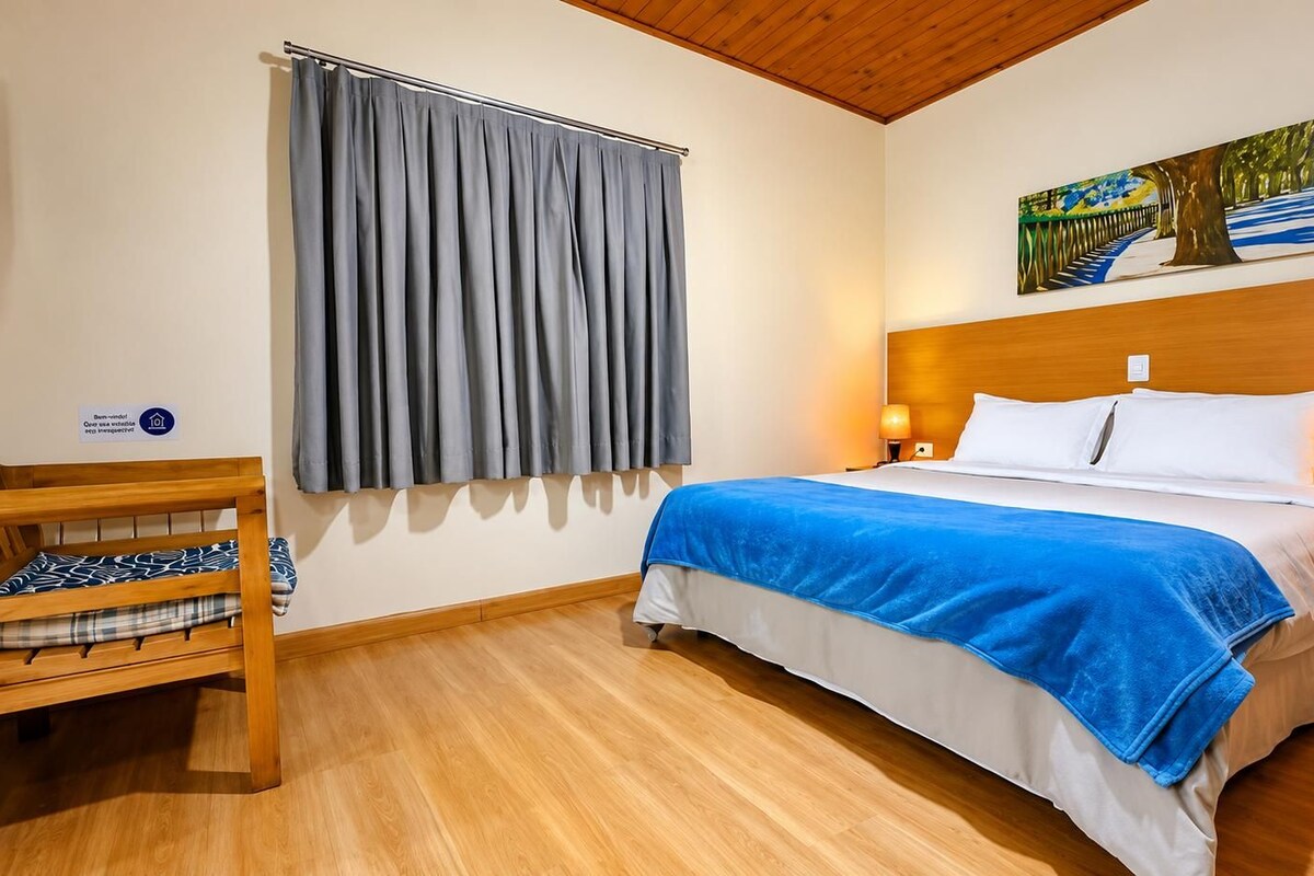 A hotel room with a bed, a wooden chair, a nightstand, a lamp, a painting on the wall, and a window with curtains.