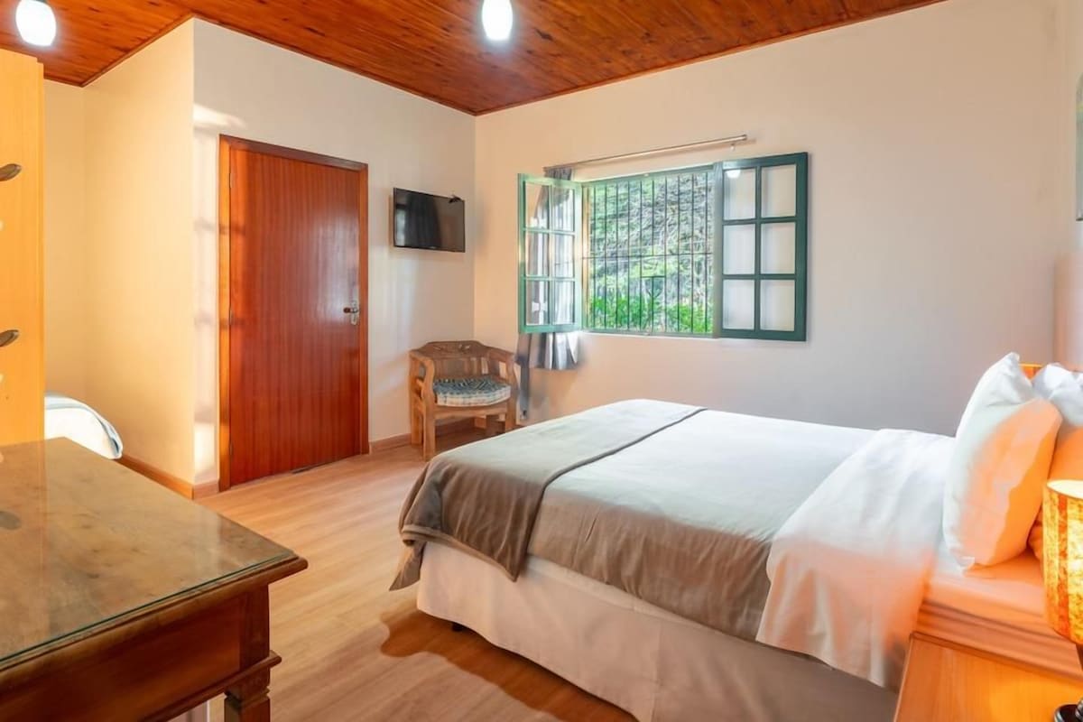 Un dormitorio con techo de madera, una cama con ropa de cama blanca, una silla, un escritorio y una ventana con vistas a zonas verdes.