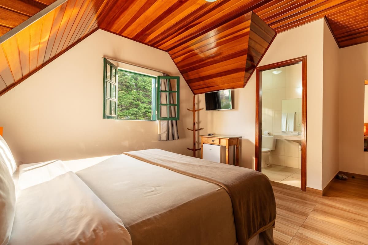 Un dormitorio con techo de madera, una cama con colcha color beige, un escritorio pequeño con espejo y vistas a un baño a través de una puerta abierta.