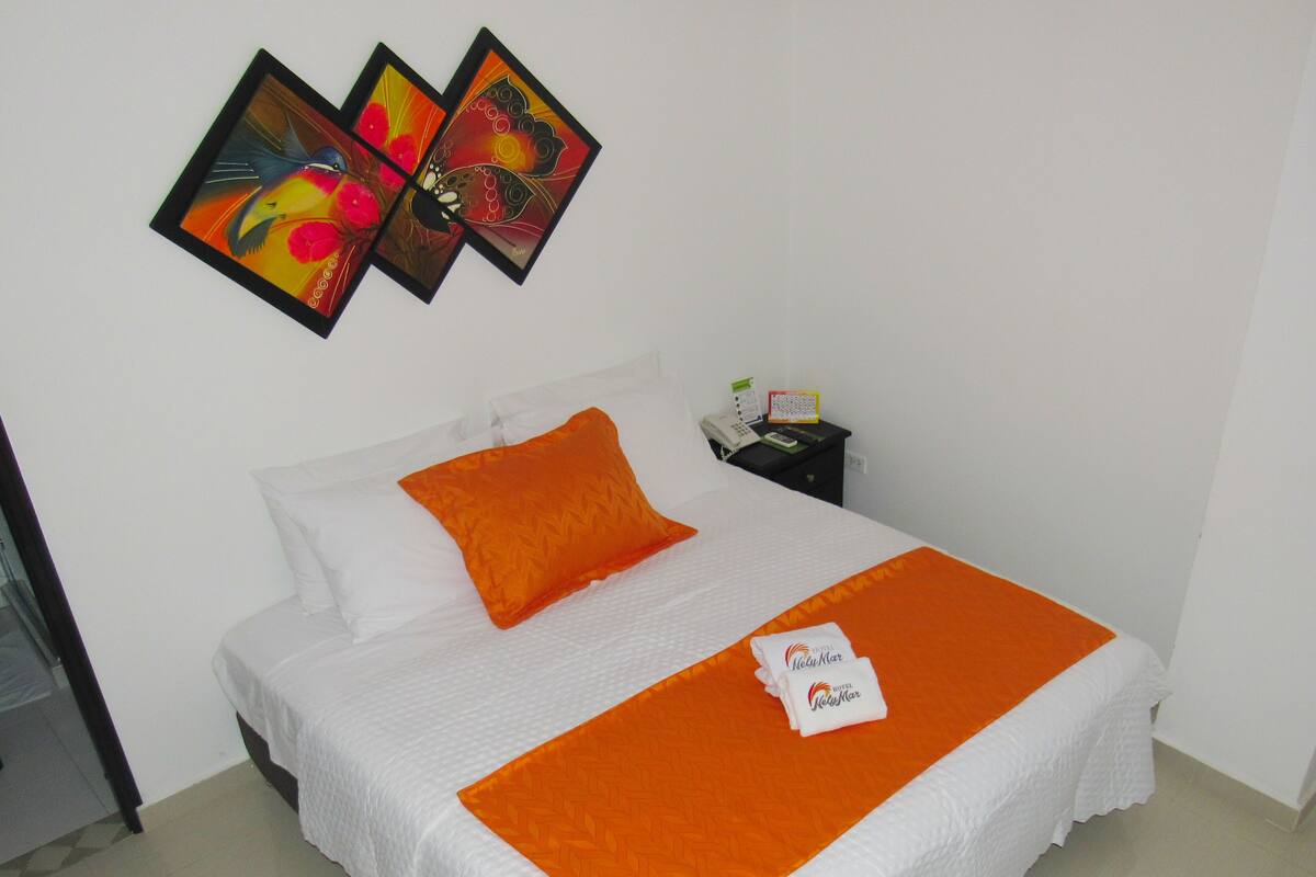 A neatly made bed with white and orange bedding, a single orange pillow, and a white towel with text. There are two framed artworks on the wall above the bed.