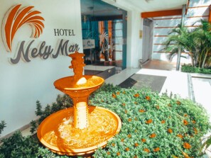 Een hotelentree met een fontein, een bord waarop "Hotel Nely Mar" staat en een tuin met goudsbloemsoorten.
