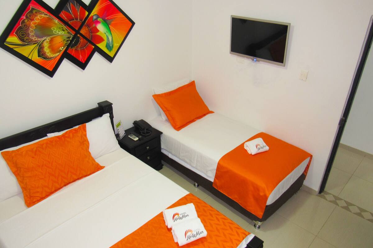 A hotel room with two beds, each with an orange bedspread and pillow, a flat-screen TV mounted on the wall, and a small table with a phone and a remote control.