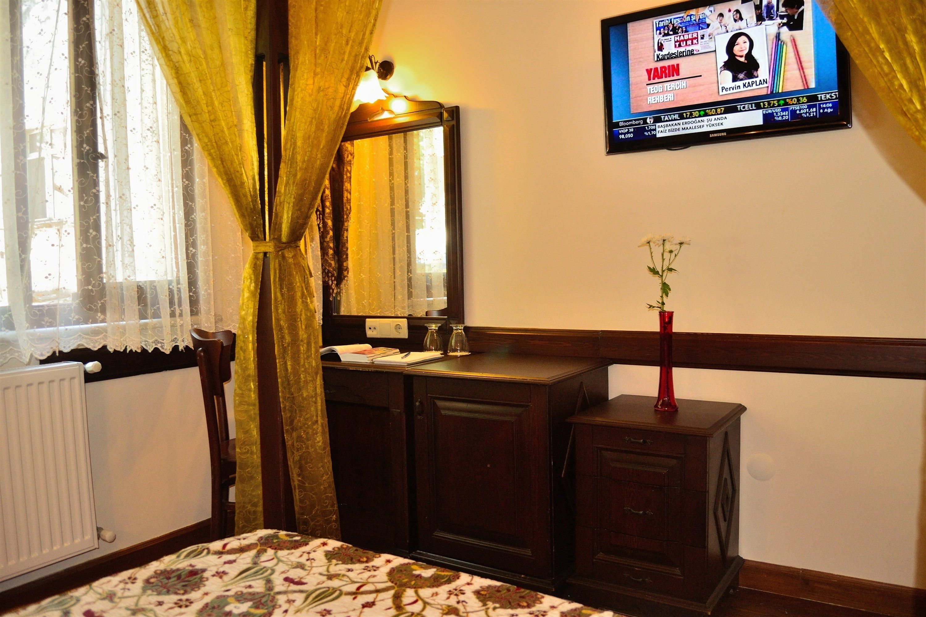 A hotel room with a bed, a wooden dresser, a mirror, and a television mounted on the wall displaying a news channel.