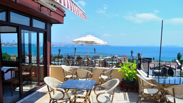 An outdoor seating area with wicker chairs and a glass table, overlooking a scenic view of the sea and city skyline. - Hotel Alp Guesthouse (Istanbul)