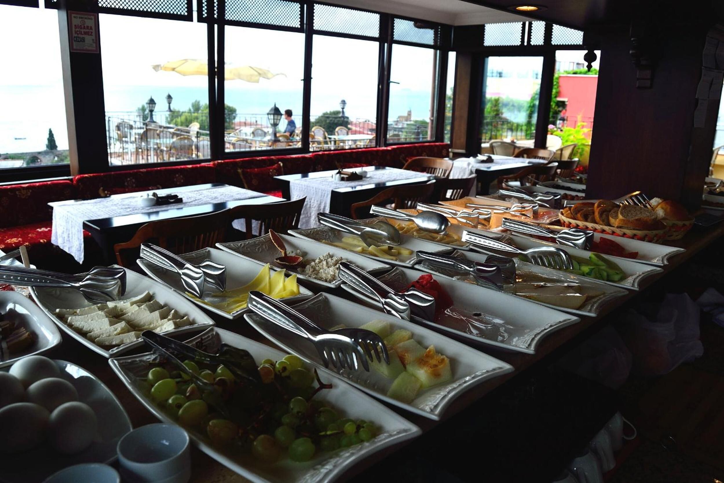 Un buffet proposant une variété de plats et d’ustensiles, des tables recouvertes de nappes blanches et une vue sur la mer par de grandes fenêtres.