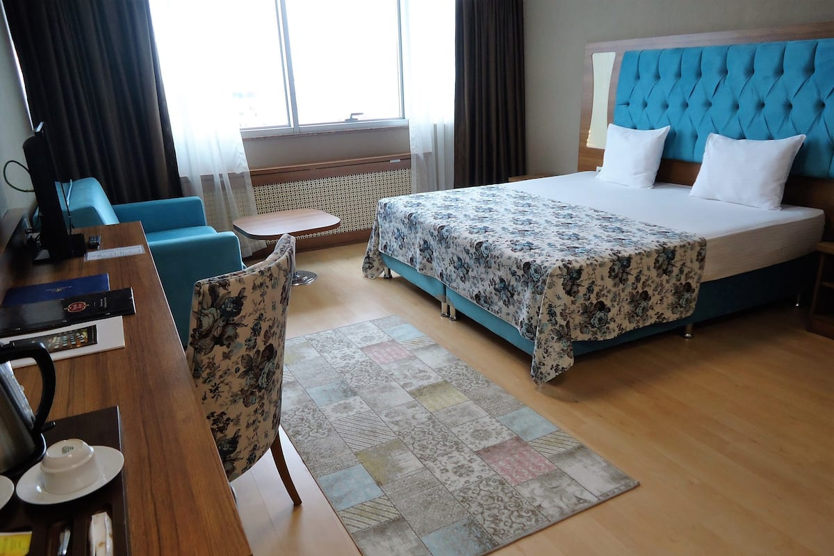 A hotel room with a bed, a desk with a chair, a small table, and a window with curtains.
