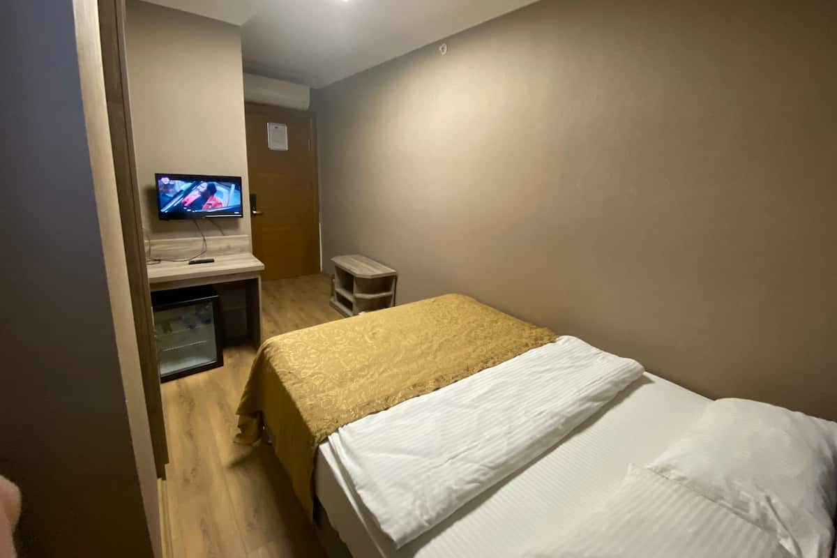 A hotel room with a bed, a desk with a television, a small stool, and a trash bin.