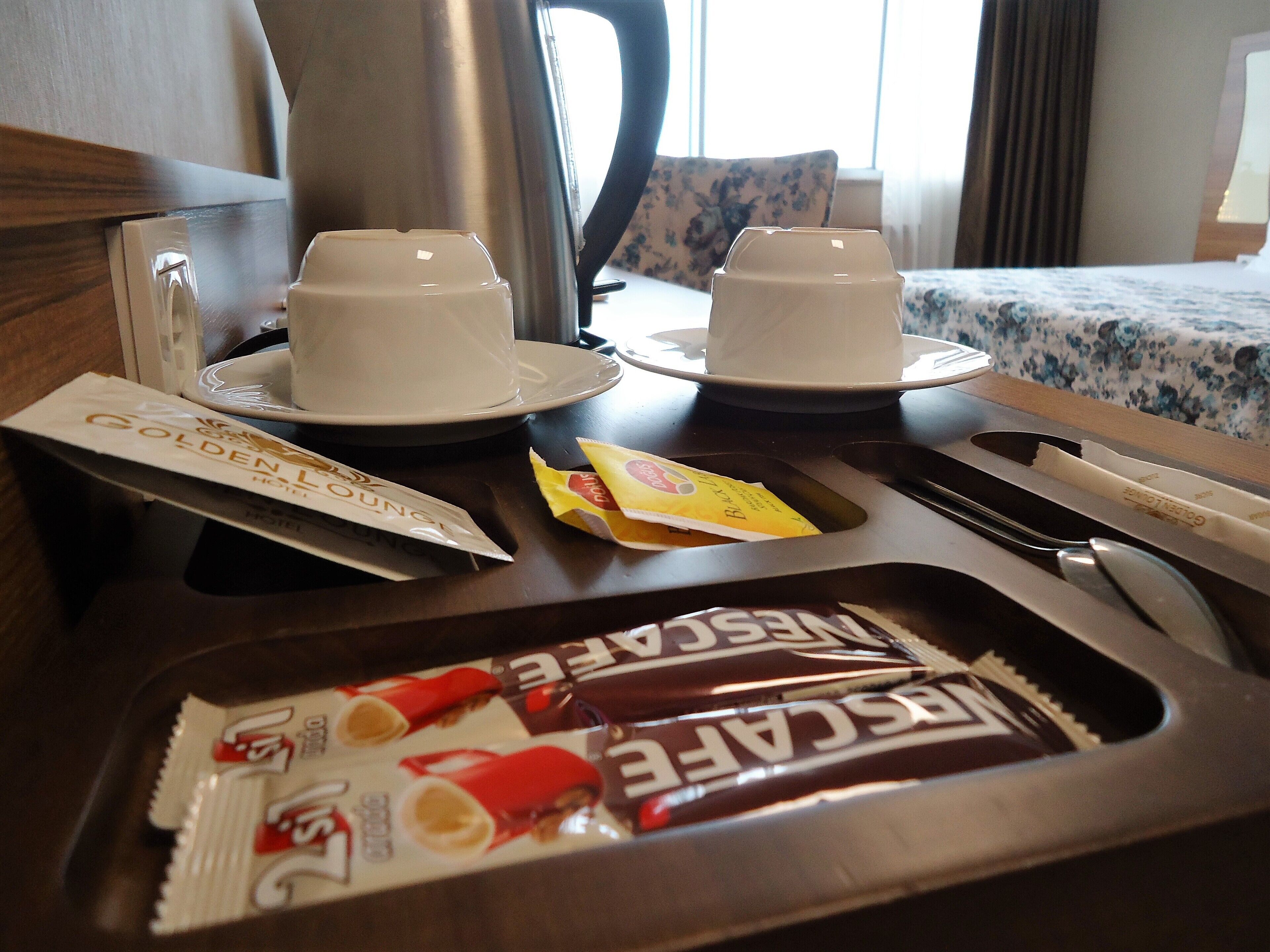 A hotel room with a bed, a coffee maker, two cups on a tray, and a tray with snacks.