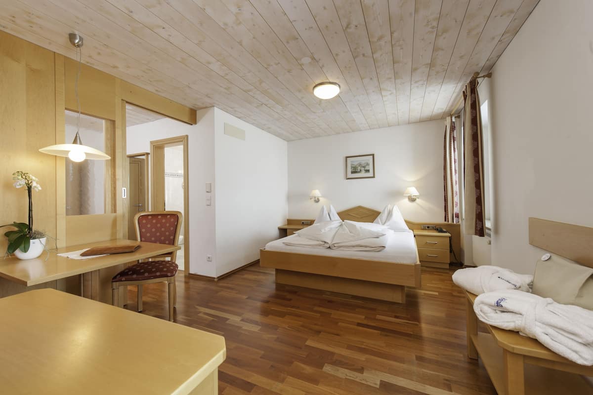 A hotel room with a wooden ceiling, a bed with white linens, a desk with a chair, a bedside table with a lamp, and a bathroom visible through an open door.