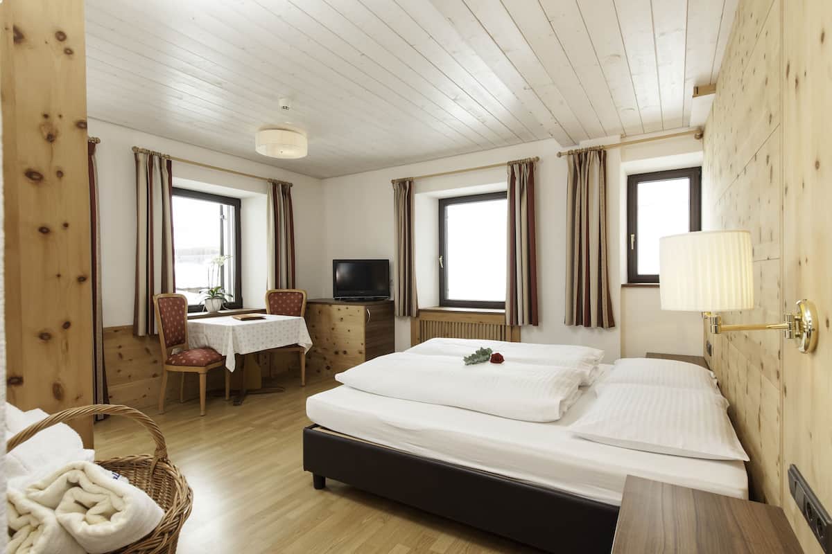 A hotel room with a wooden ceiling, a bed with white bedding, a television, a dining table with chairs, and a window with curtains.