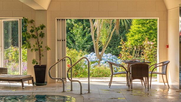 Zona de piscina con una silla de salón, una mesa con sillas y vistas a un jardín a través de amplios ventanales.