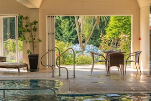 Zona de piscina con una silla de salón, una mesa con sillas y vistas a un jardín a través de amplios ventanales.
