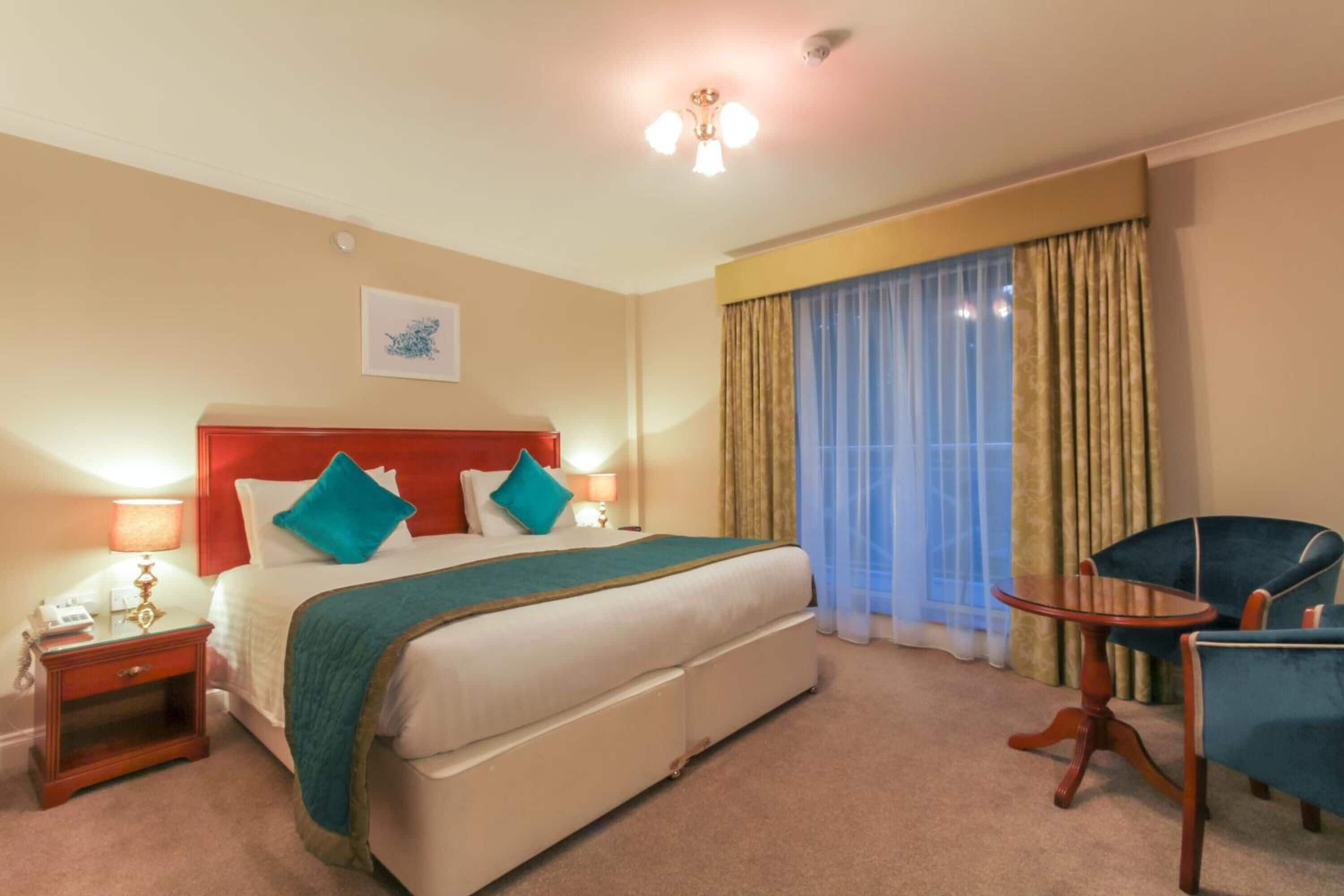 A hotel room with a large bed, a bedside table with a lamp, a chair, and a window with curtains.