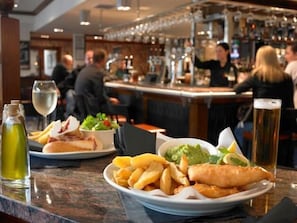 Zona de bar con mostrador, vasos de cerveza y un plato de comida que incluye pescado con papas fritas.