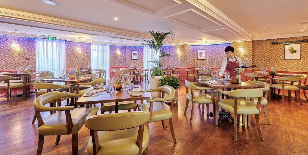 Un comedor de hotel con varias mesas y sillas, un camarero atendiendo a los huéspedes y una pared decorativa con un diseño en zigzag.