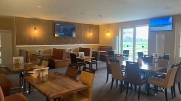 A hotel dining area with wooden tables and chairs, a large TV screen on the wall, and a view of the outdoors through large windows.