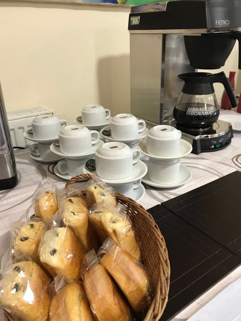 A coffee machine, multiple white cups and saucers, and a basket of pastries on a table.