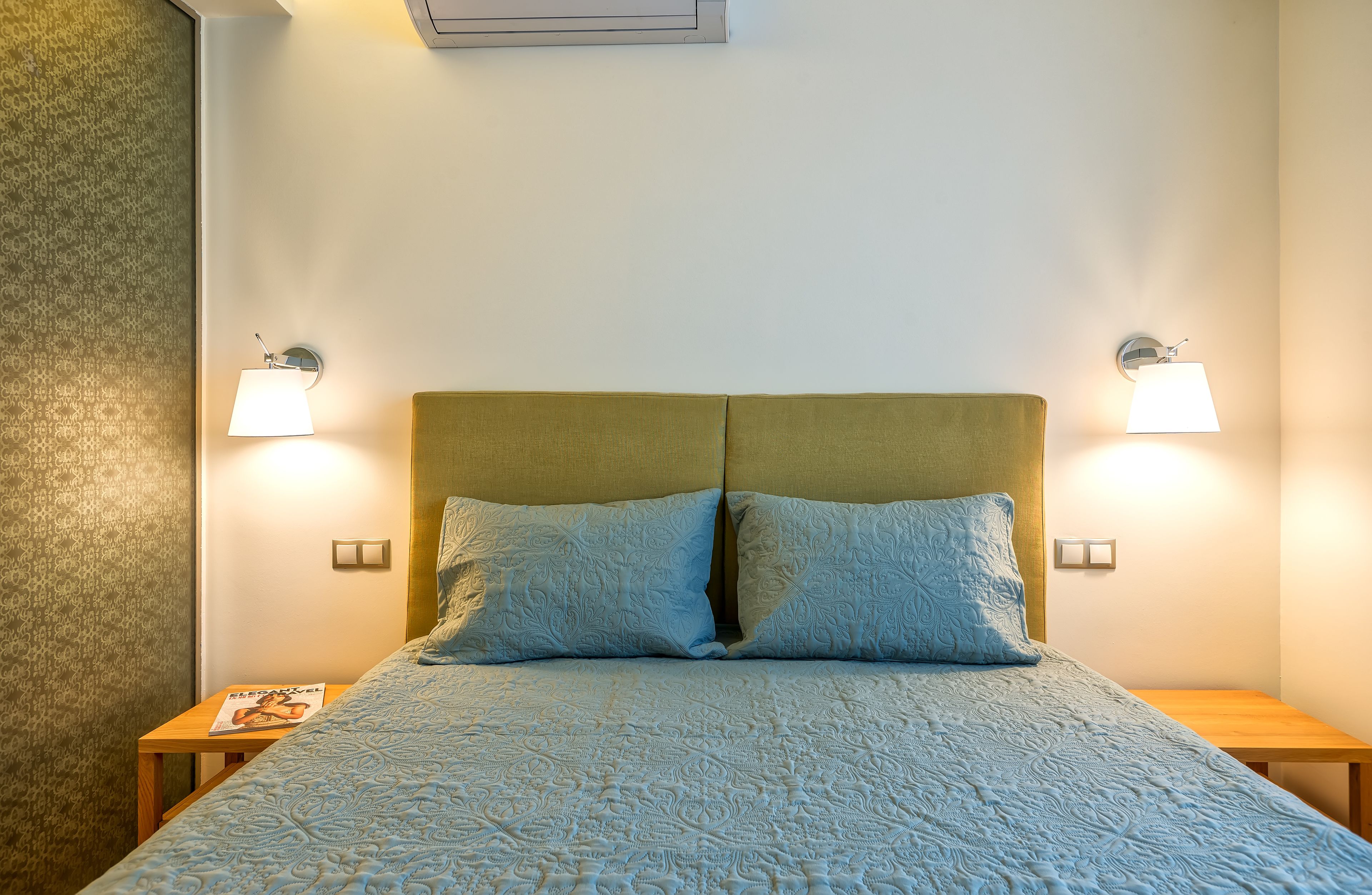 A neatly made bed with a patterned bedspread, two wall-mounted lamps, and a small wooden nightstand with a magazine.