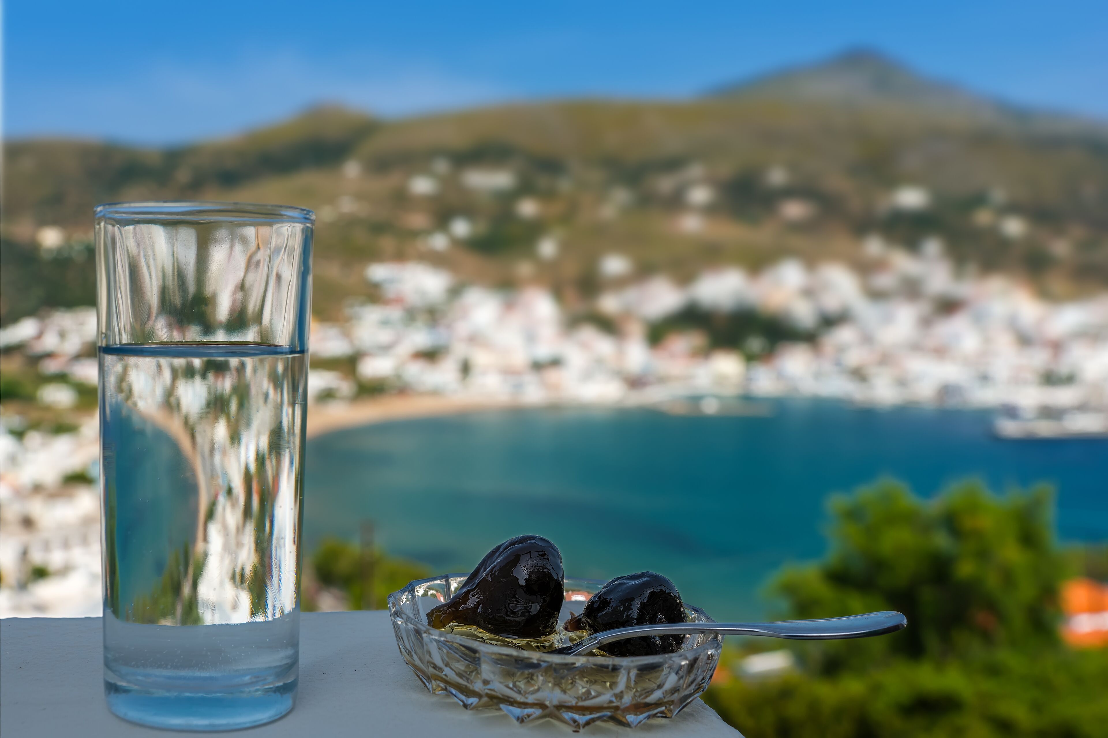 A glass of water and two olives on a table with a scenic view of a coastal town and mountains in the background.