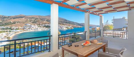A balcony with a wooden table and wicker chairs, overlooking a coastal town and a bay.