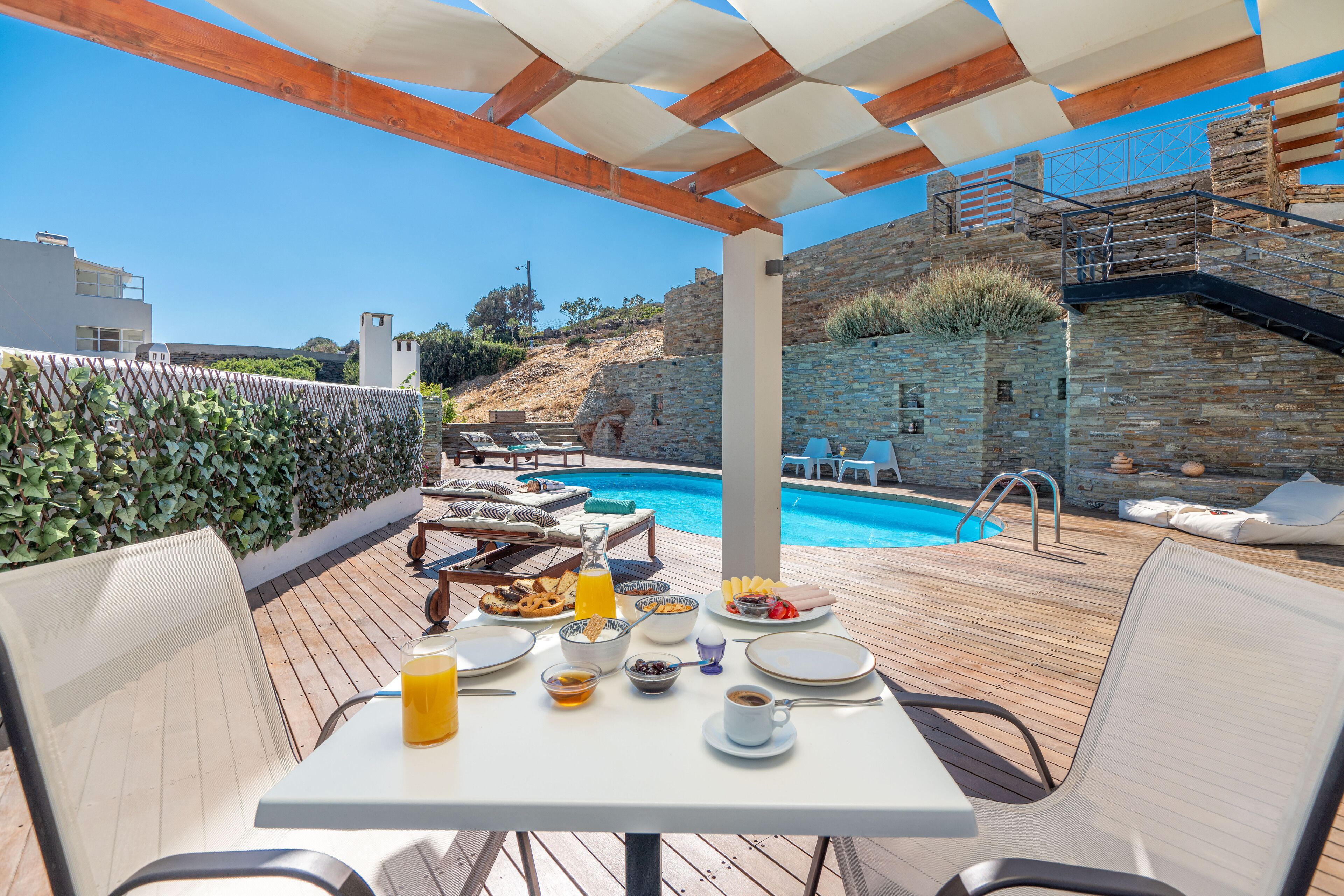A dining table set for breakfast with a pool view, wooden deck, and stone walls.