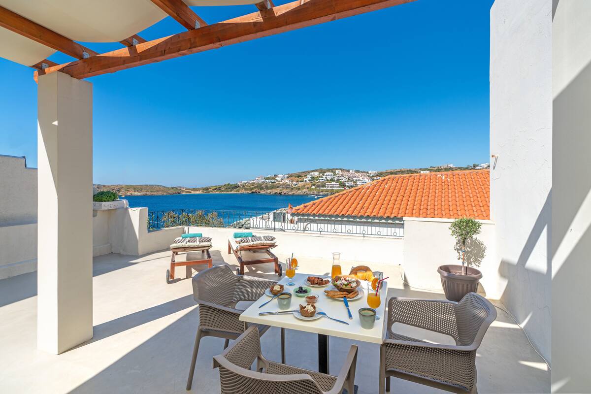 A terrace with a table set for breakfast, overlooking a coastal town.