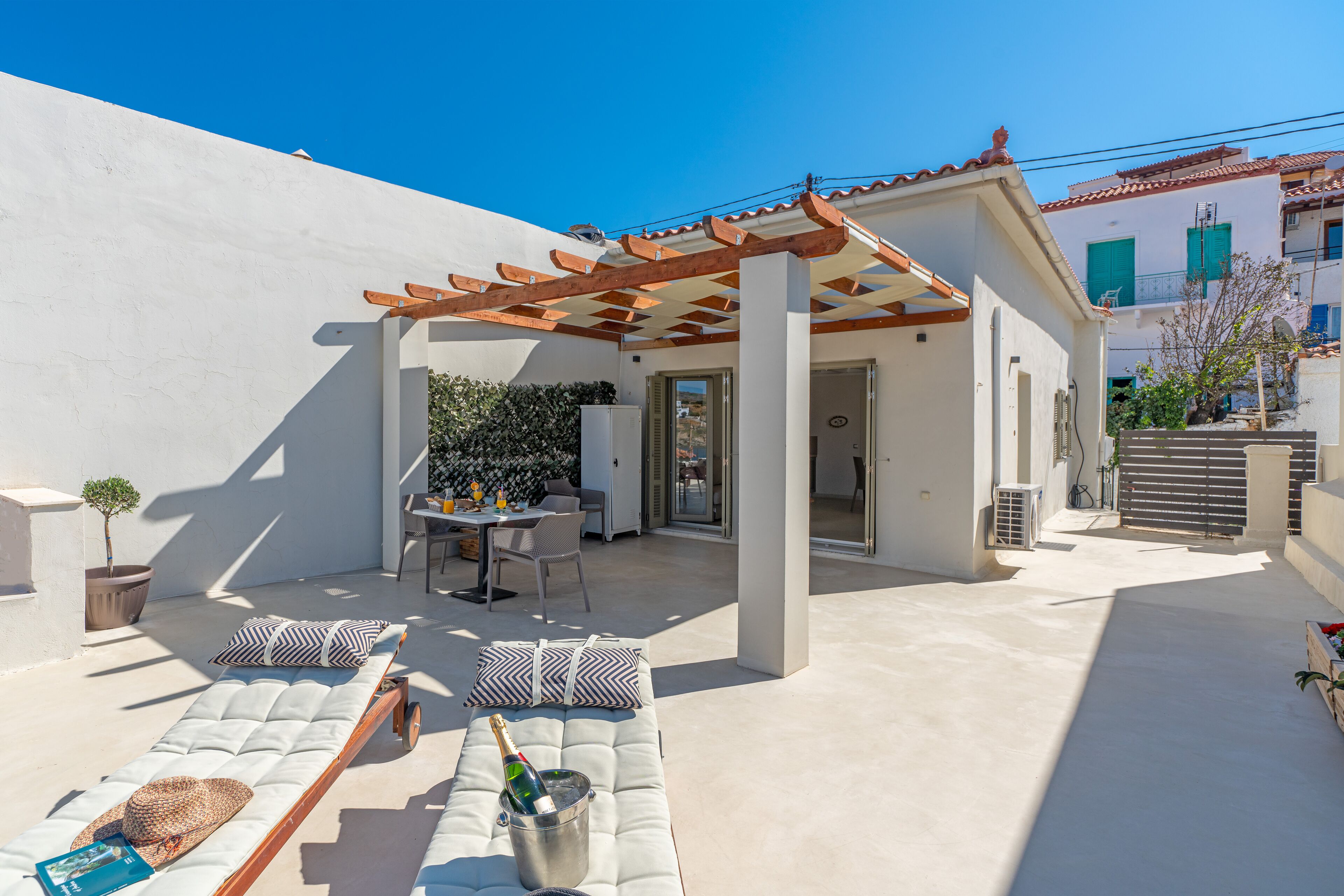 Un espacio al aire libre con sol, un pérgola, sillones cómodos con cojines y una mesa con sillas.