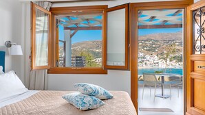 A bedroom with a bed, two pillows, a nightstand, a lamp, and a window with a view of a coastal town. - Belvedere Andros (Andros)
